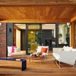 A stylish covered outdoor patio features modern white lounge seating with colorful pillows, a wooden coffee table with drinks and snacks, and a sleek navy bench. White Sonos outdoor speakers are installed on the roof.